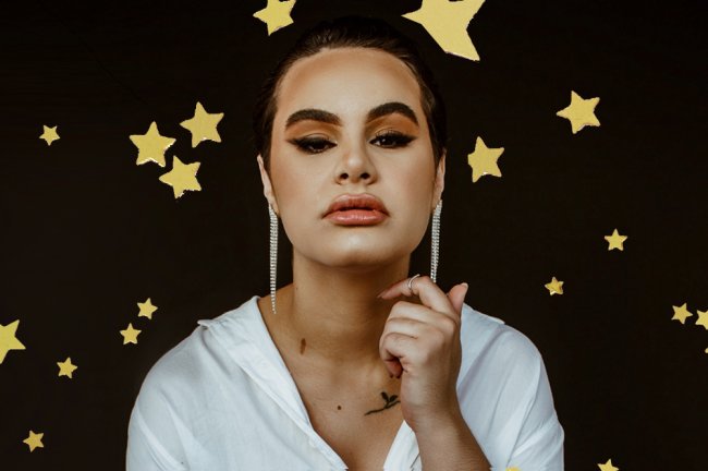 A moda que você veste é uma imposição ou realmente expressa quem você é? Imagem de uma mulher olhando para a câmera. A mão está próxima do rosto e ela está com uma make preta carregada.