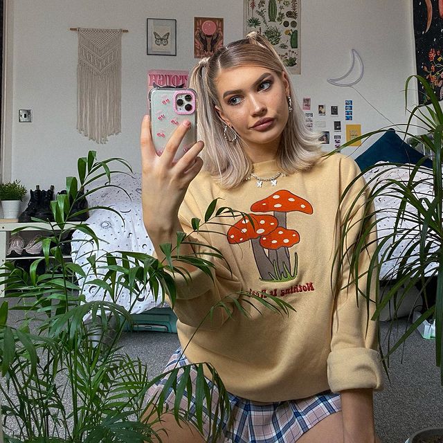 moletom cogumelo Selfie no espelho de uma mulher no quarto. Ela usa um moletom com estampa de cogumelo, saia xadrez e cabelo preso em uma maria-chiquinha. Ela olha para a câmera e não sorri.