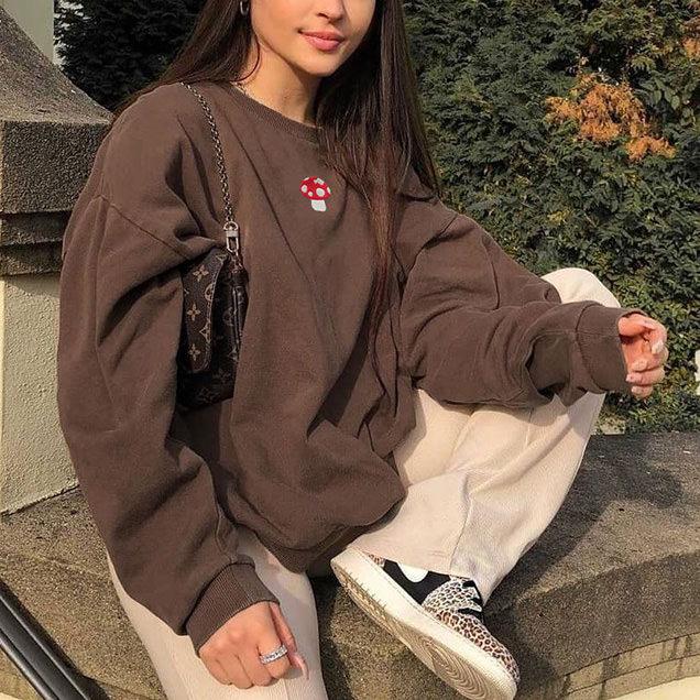moletom estampa cogumelo Foto de uma mulher sentada em um muro. Ela usa um moletom marrom com estampa de cogumelo, calça baggy cor creme, tênis e bolsa baguete. Ela olha para a câmera e sorri.