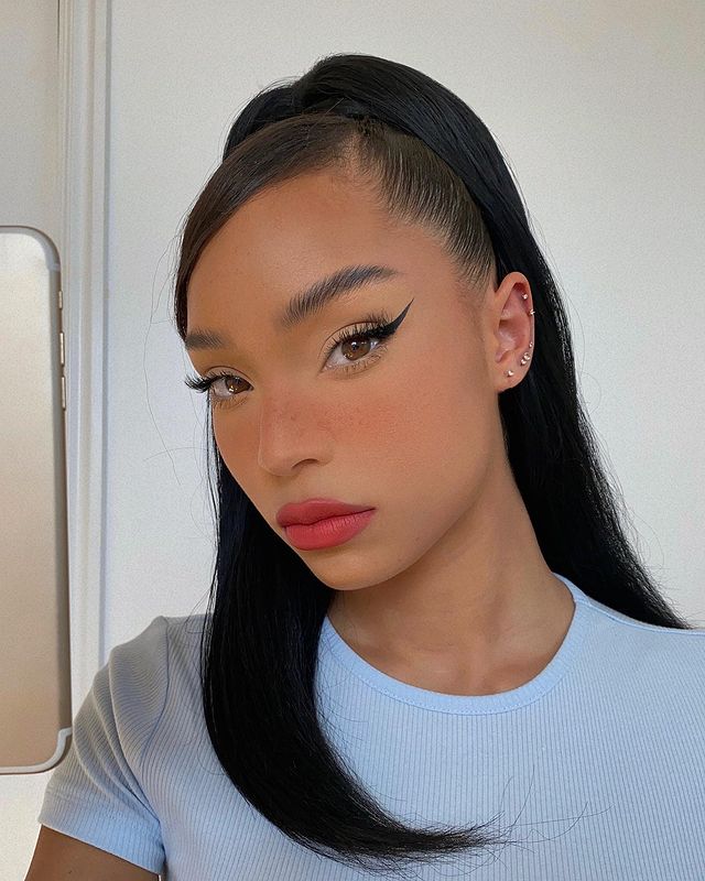 penteado rabo de cavalo Selfie de uma mulher com o penteado rabo de cavalo alto. Ela usa uma blusa canelada azul e maquiagem básica com delineado. Ela olha para a câmera e não sorri.