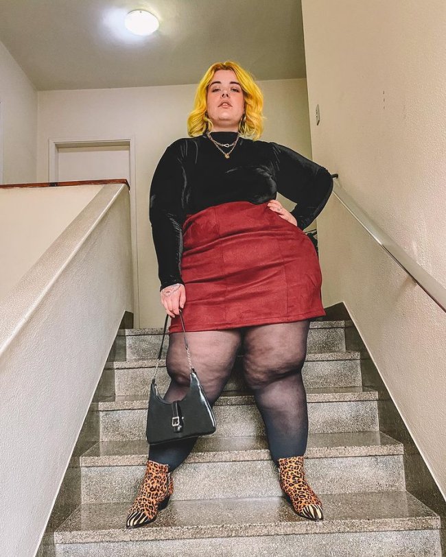 saia com blusa de gola alta Foto de uma mulher em uma escada. Ela usa uma blusa preta de gola alta, saia de couro vermelha, meia calça, bota com estampa animal print e bolsa preta. Ela olha para a câmera e não sorri.