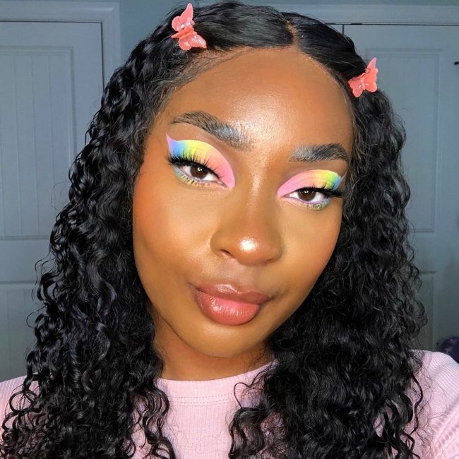sombra colorida tons pastel Selfie de uma mulher. Ela usa uma camiseta rosa, cabelo solto com acessório em formato de borboleta na franja, maquiagem com pele natural e sombra colorida em tons pastel. Ela olha para a câmera e sorri levemente.