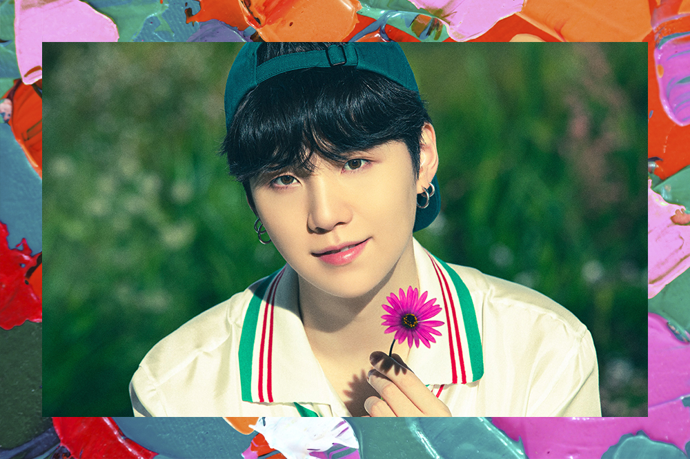 Suga, do BTS, está de boné segurando uma flor rosa. Ao fundo há vegetação