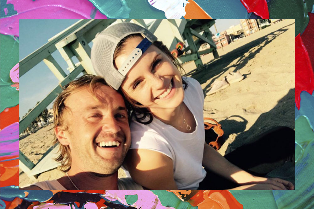 Na imagem o ator Tom Felton está sorridente encostando a cabeça na de Emma Watson ao lado, também sorrindo. Os dois estão na praia.