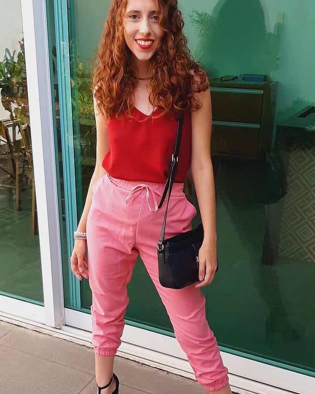 Bolsa preta quebrando o visual Jovem posando com regata vermelha, calça rosa e bolsa preta. Sua expressão é sorridente.