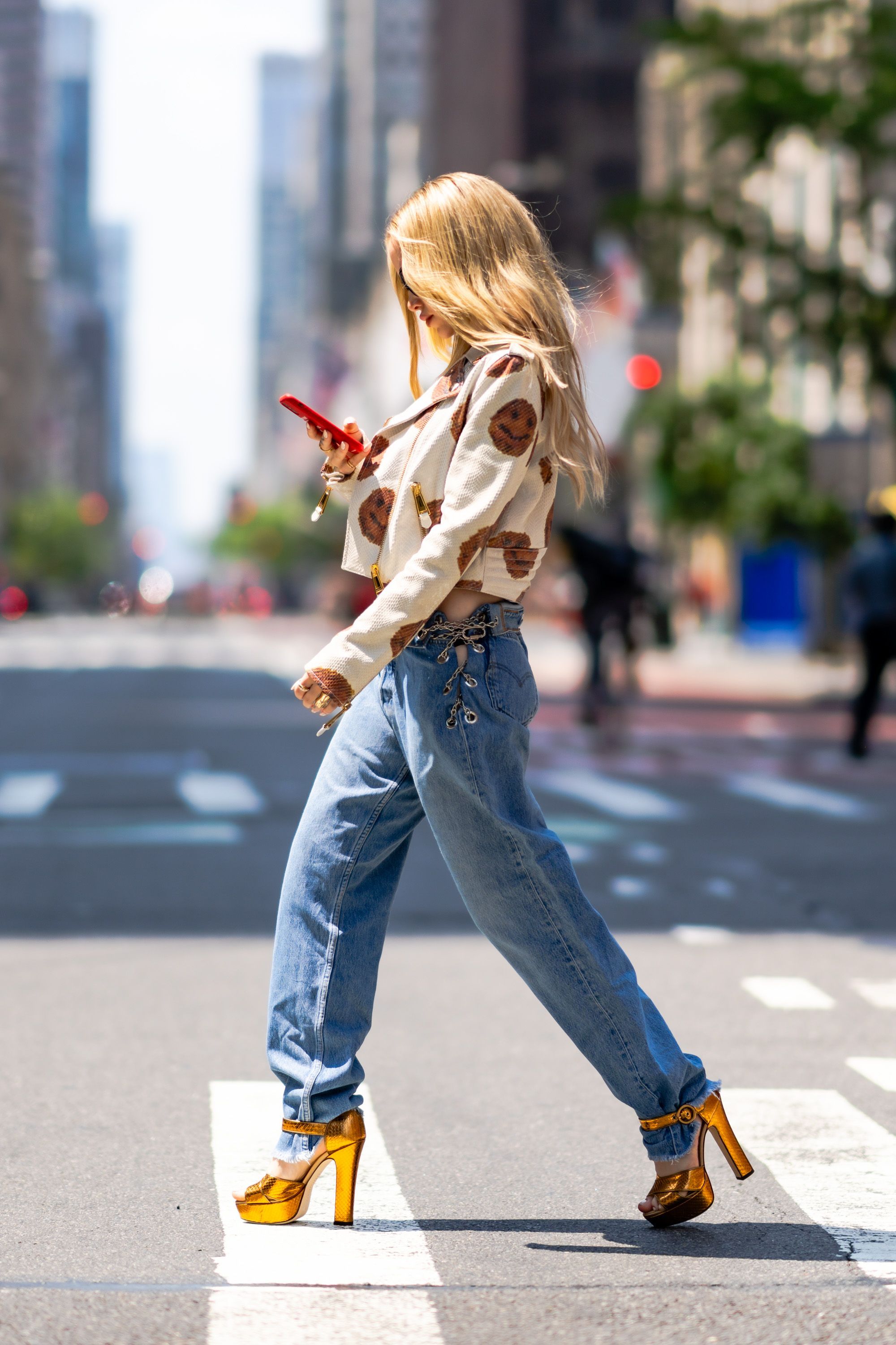 Dove Cameron usando calça com cintura baixa Foto da atriz e cantora Dove Cameron de lado, caminhando em uma rua. Ela está olhando para seu celular, usando jaqueta e calça jeans.