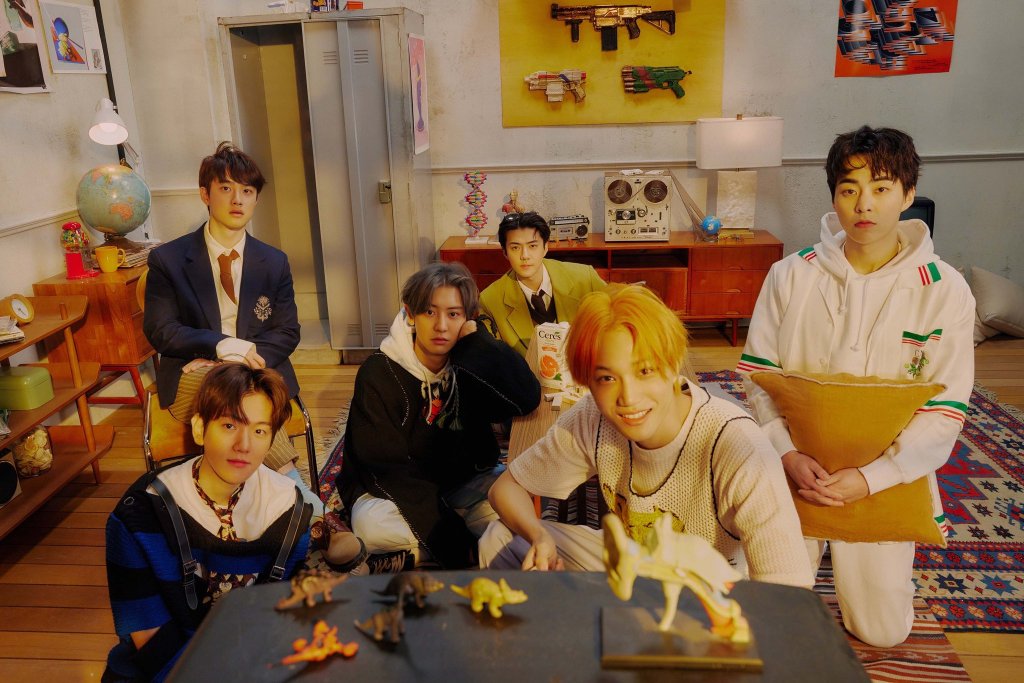 Imagem de divulgação do grupo EXO posando em um quarto com objetivos cheios de detalhes em tons de amarelos; assim como a iluminação do ambiente; uma mesa com miniaturas de dinossauros aparece em primeiro plano e os integrantes em segundo