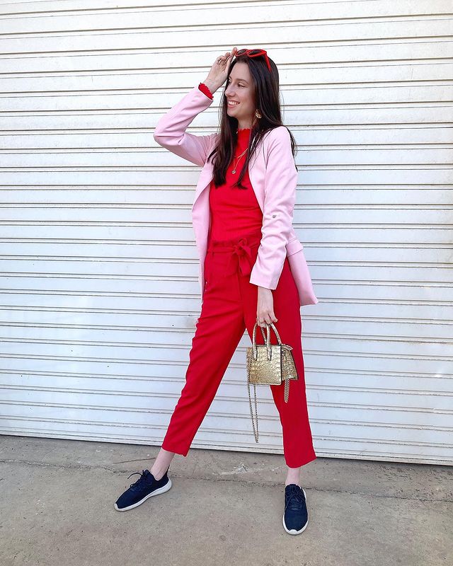 Look vermelho e rosa Jovem posando em frente a parede branca com blazer rosa claro, uma das mãos apoiadas na cabeça e segurando uma bolsinha com tênis preto.