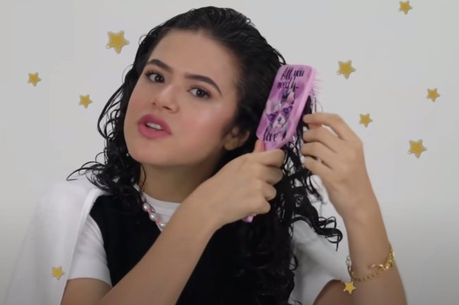 Maisa Silva Print de vídeo do youtube no qual Maisa finaliza seu cabelo. Ela usa blusa branca com suéter preto, penteia seu cabelo com escova branca e usa colar de miçanga.