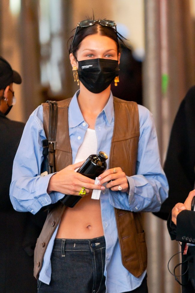 bella-hadid-colete-de-couro Foto da modelo Bella Hadid na rua. Ela usa um top cropped branco, camisa azul aberta, colete de couro marrom, calça jeans escura, máscara facial preta, óculos de sol no topo da cabeça e cabelo preso em um coque. Ela olha para a frente e não sorri para a foto.