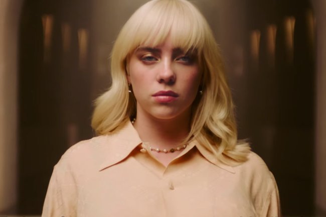 billie eilish Billie Eilish usando camisa amarela, com expressão facial séria olhando para a câmera. Está de cabelo médio e franjinha em loiro platinado