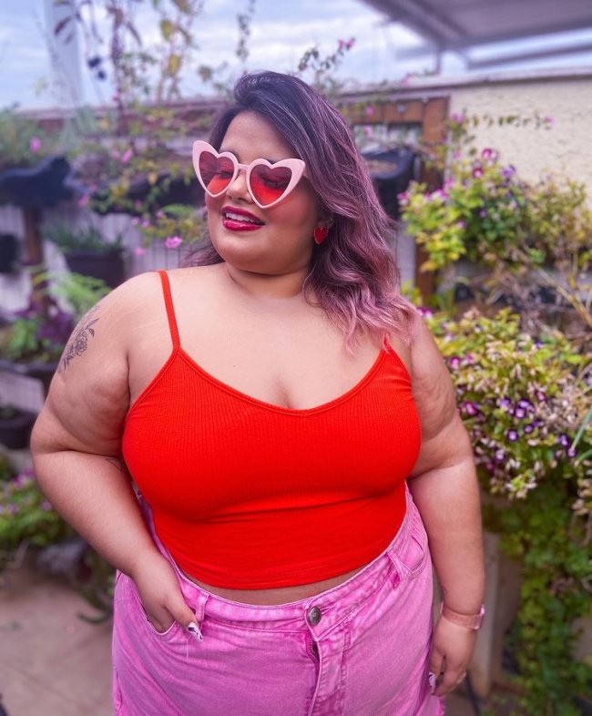 blusa vermelha e saia rosa Jovem sorridente posando com óculos de coração, regata vermelho e calça jeans rosa.