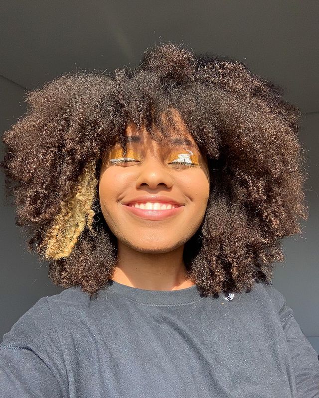 Selfie de uma mulher sorrindo. Ela usa uma camiseta preta e cabelo crespo solto.