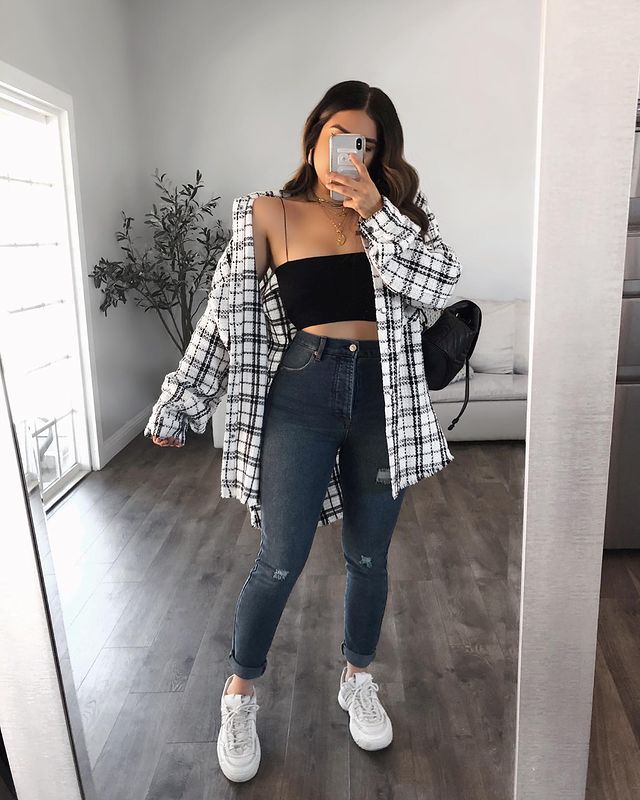 Selfie no espelho de uma mulher mostrando o look. Ela usa uma calça skinny escura, cropped preto, camisa xadrez oversized branco e preta, bolsa preta e tênis branco.