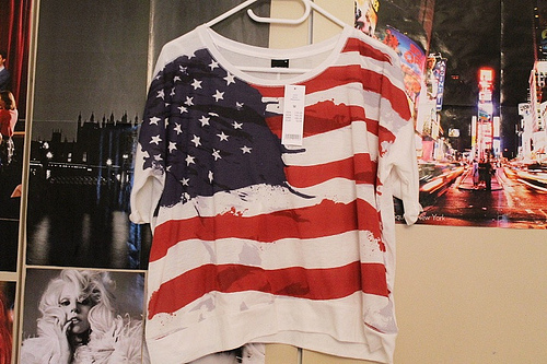 camiseta-bandeira-eua Foto de uma camiseta com estampa dos Estados Unidos pendurada em um cabide na parede de um quarto.