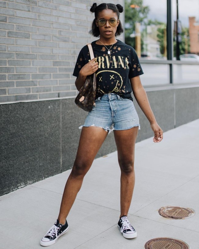 camiseta de banda com short e all star Jovem posando com short jeans e camiseta de banda. Sua expressão é séria e ela usa o penteado space buns em seus cabelos.