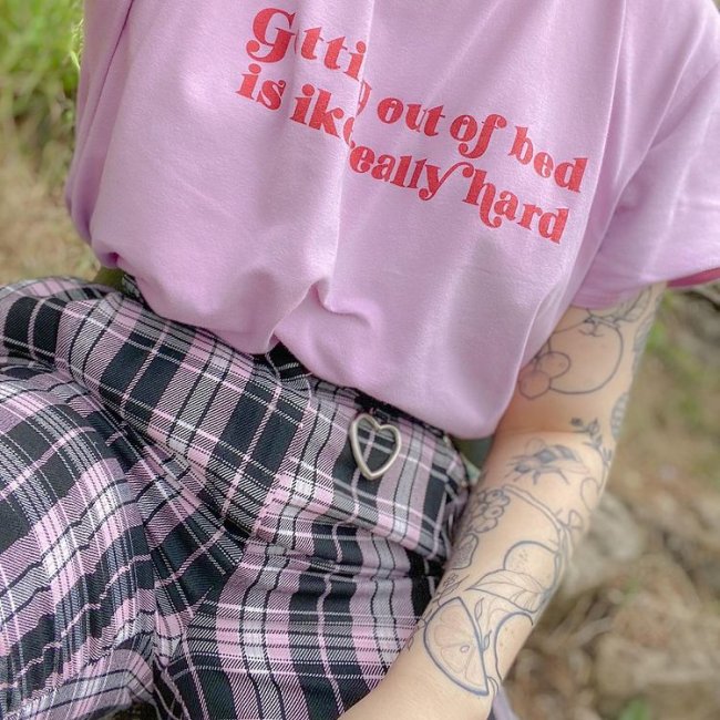 camiseta rosa e vermelhjja Na imagem só podemos ver a camiseta rosa com uma frase escrita em vermelho. A pessoa também uma calça xadrez lilás e preta.