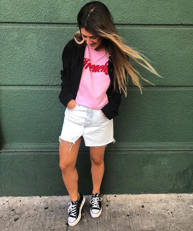 camiseta rosa Jovem posando em porta verde enquanto olha para baixo, ela usa casaco preto, saia branca e camiseta rosa com palavra em vermelho.