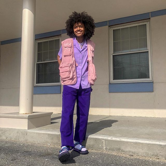 colete-cargo-rosa Foto de uma mulher em uma escada. Ela usa uma camiseta lilás, colete cargo rosa claro, calça azul, meia e papete. Ela está com as mãos nos bolsos do colete, olha para frente e sorri.