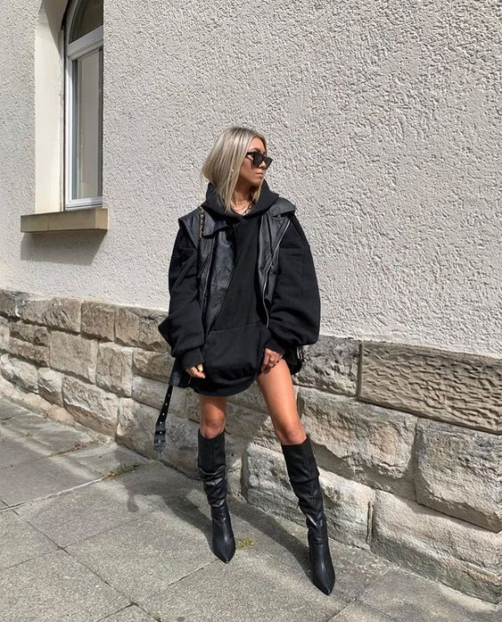 colete-de-couro-look Foto de uma mulher na rua. Ela usa uma blusa de moletom preta oversized como vestido, bota preta modelo over the knee, bolsa preta, colete de couro preto como sobreposição, óculos escuros e cabelo solto. Ela olha para o lado e não sorri.