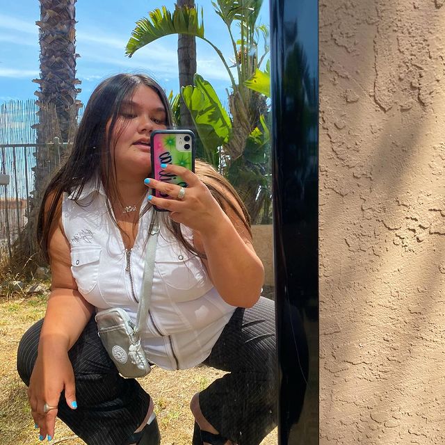 colete-jeans-look Selfie no espelho de uma mulher. Ela usa uma calça alfaiataria escura, colete jeans claro fechado, bolsa branca e está com o cabelo liso e longo solto. Ela está agachada, olha para o celular e não sorri.