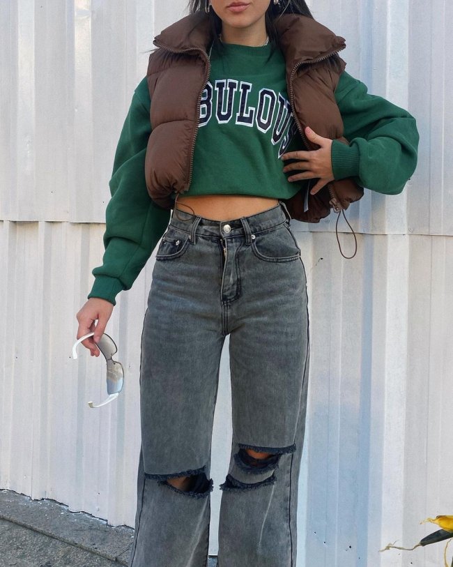 colete puffer look Foto do look de uma garota. Ela usa uma blusa de moletom verde escuro, um colete puffer marrom, calça modelo wide leg cinza. Ela está com a mão esquerda no colete e segura um óculos de sol com a mão direita.