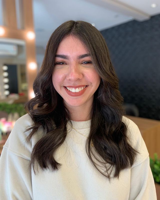 corte em camadas cabelo castanho Jovem sorridente usando camisa branca e posando em salão de cabeleireiro.