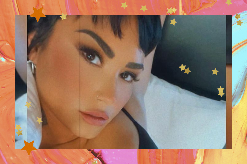 Selfie de Demi Lovato; ela está olhado para câmera com expressão séria e usando um sutiã preto; a margem é uma textura de tintas em tons de laranja, amarelo, rosa, verde e estrelas amarelas e laranjas decoram a imagem