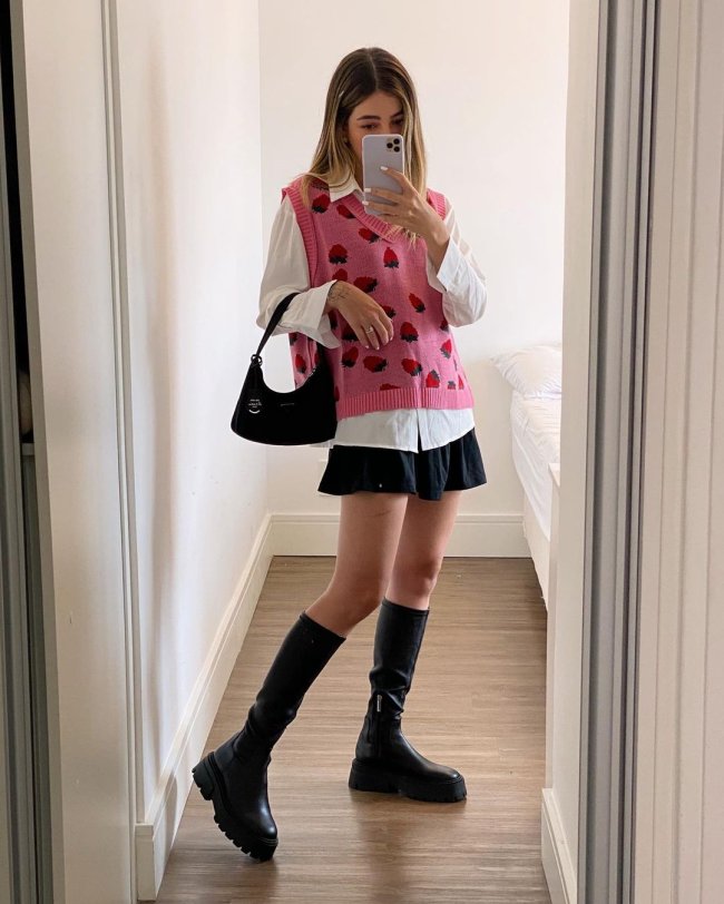 look-colete-de-vovo-rosa Selfie no espelho de uma mulher. Ela usa uma camisa branca, colete de vovô rosa com estampa de morangos como sobreposição, saia preta, coturno preto e bolsa preta. Ela olha para a tela do celular e não sorri.