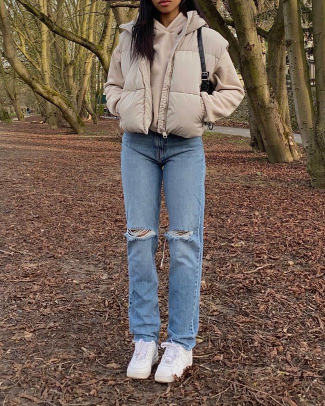 look-colete-puffer Foto do look de uma garota. Ela usa uma blusa de moletom bege, colete puffer bege, calça jeans e tênis branco. Ela está com as duas mãos no bolso do colete e não é possível ver o rosto dela na foto.