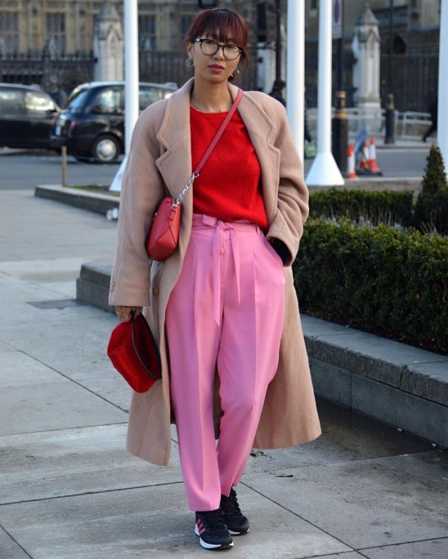 look rosa e vermelho com quebra de cor Jovem posando para foto na rua usando blazer bege e camisa vermelha com calça rosa, ela está com uma das mãos no bolso e a outra segura uma bolsa vermelha.