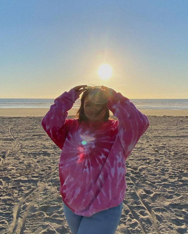 look-tie-dye-rosa Foto de uma mulher na praia. Ela usa um moletom tie dye rosa, calça jeans e óculos de sol na cabeça. Ela segura o óculos de sol no topo da cabeça com as duas mãos, olha para a câmera e sorri levemente.