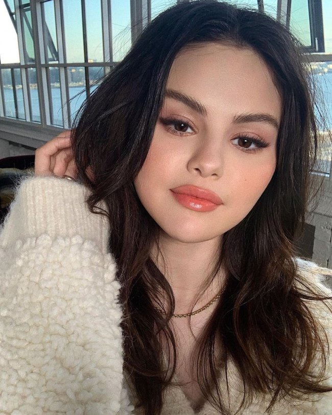 maquiagem-selena-gomez Selfie da atriz e cantora Selena Gomez. Ela usa um casaco bege de pelinhos, cabelo solto, olho esfumado, gloss e uma corrente dourada. Ela olha para a câmera, sorri levemente e está com a mão direita perto do rosto.