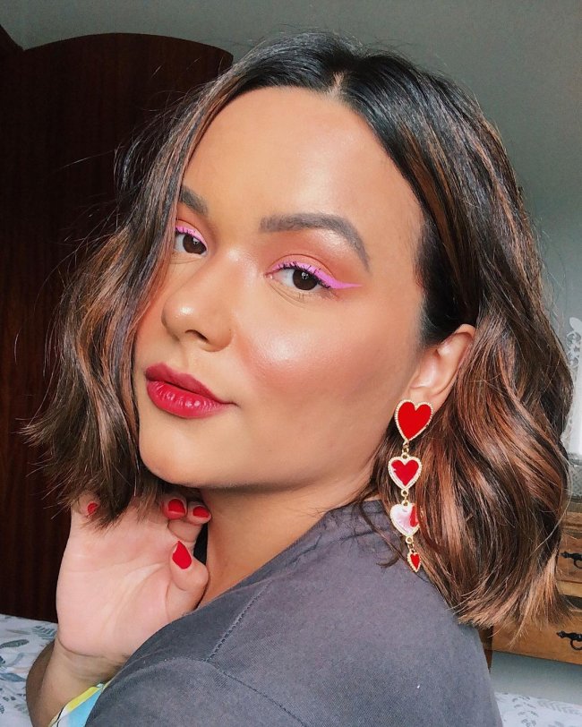 maquiagem Jovem posando para selfie com expressão séria, ela usa delineador rosa e brincos em formato de coração vermelhos.