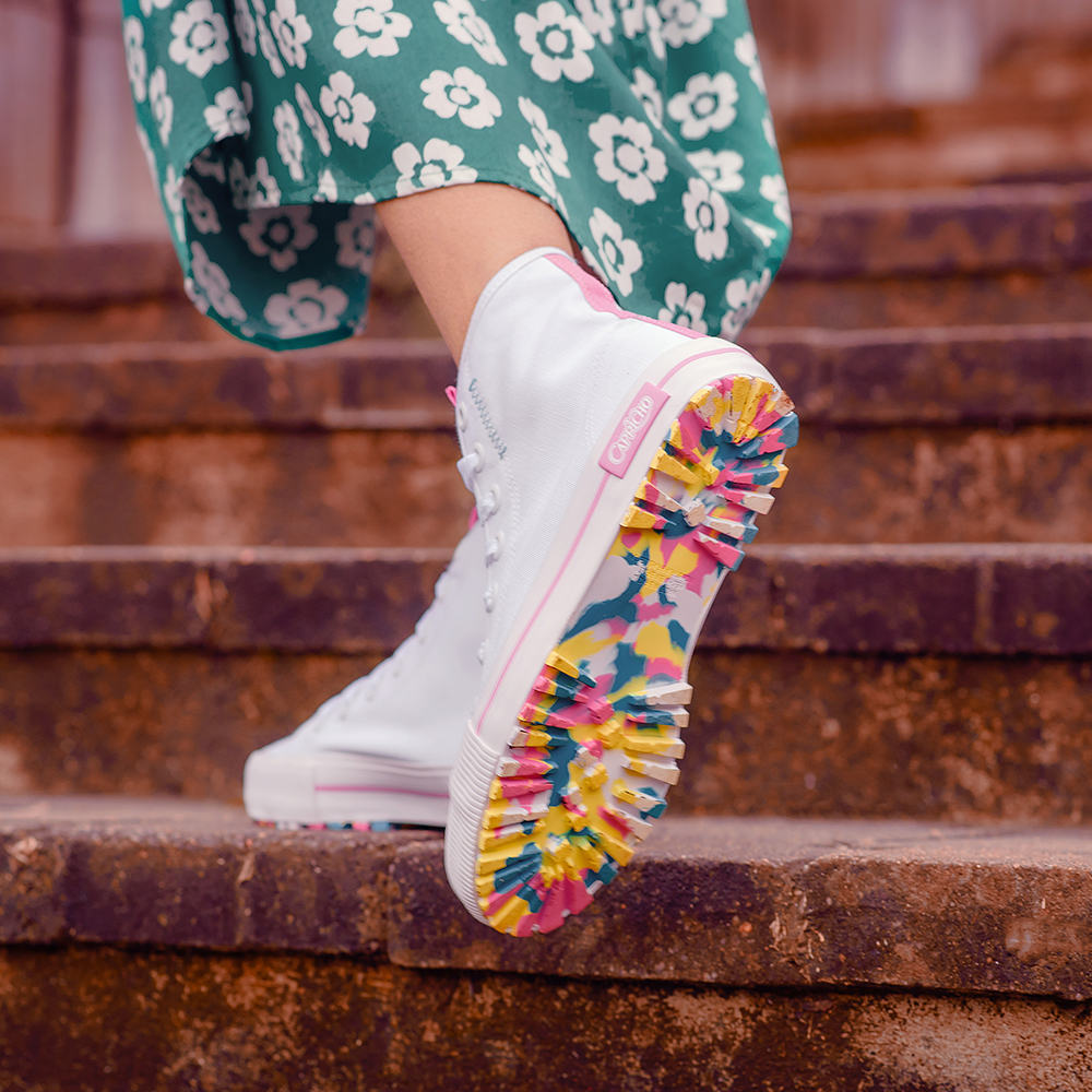 nova-colecao-tenis-capricho-shoes-02 Foto do pé de uma menina usando tênis branco de cano alto da CAPRICHO Shoes com sola toda colorida. Ela está em um degrau de uma escada e dá para ver um pedaço do seu vestido longo verde com flores.