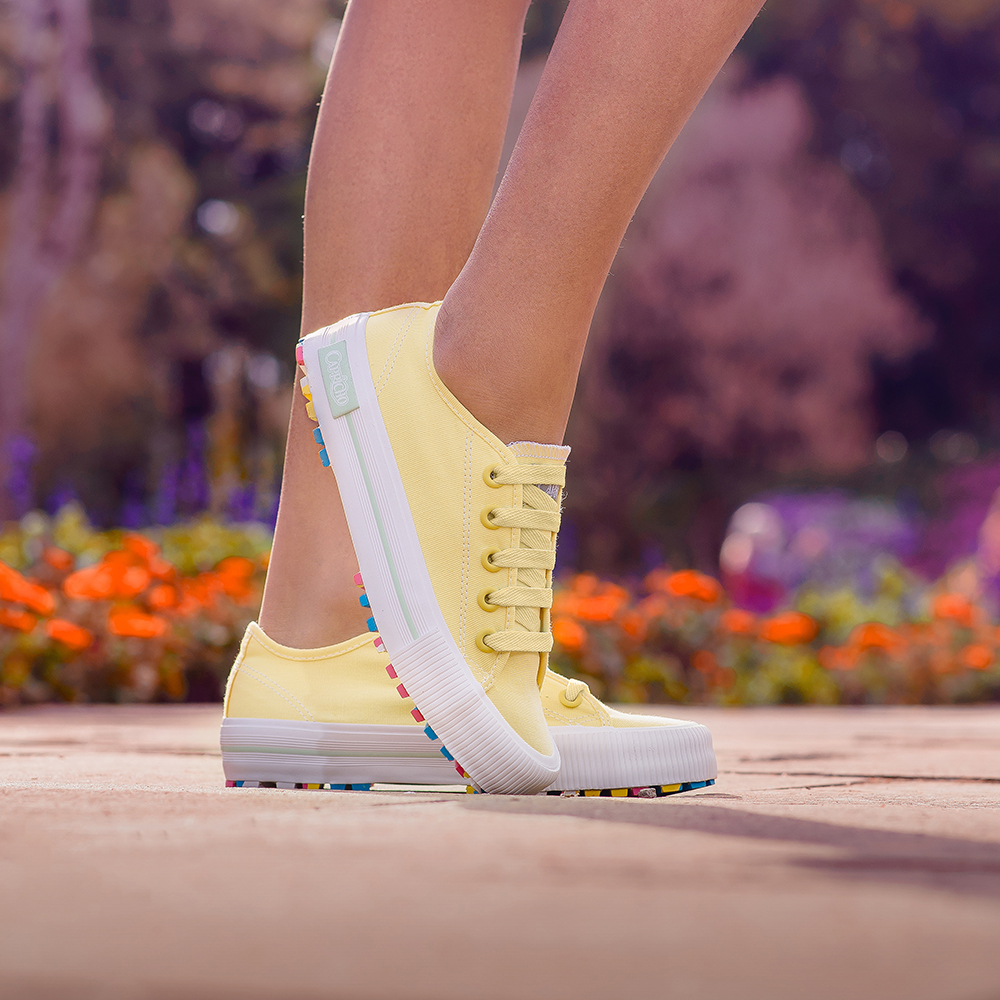 Garota usando tênis amarelo com sola colorida da coleção da CAPRICHO Shoes. Na foto, dá para ver os pés dela no chão, e um deles está na frente com a ponta do tênis tocando o chão.