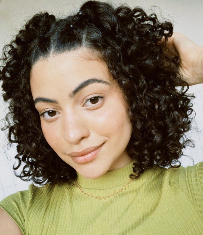 penteado cabelo cacheado curto Jovem sorridente posando para selfie usando camisa verde com gola alta, e posando com uma das mãos apoiadas na cabeça.