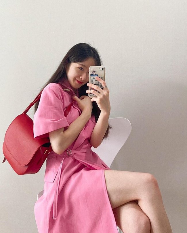 roupa com tom preto e rosa Jovem sentada em uma cadeira branca, ela aponta o celular para o espelho e seu look é composto por vestido rosa e uma bolsa vermelha que está pendurada em seu braço esquerdo.