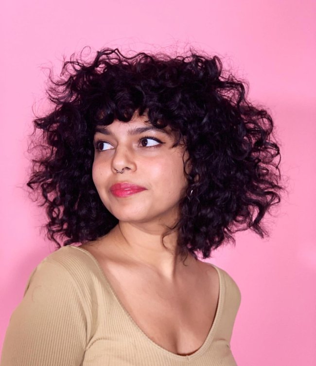 Foto de uma mulher sorrindo e olhando para o lado. Ela usa uma blusa bege e cabelo solto no corte shaggy.