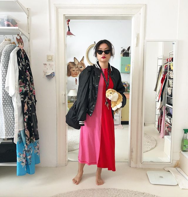 vestido vermelho e rosa Mulher posando frente a espelho do quarto, podemos ver alguns detalhes do cômodo como as cores brancas da parede e uma arara com roupas. Ela usa vestido metade vermelha e metade rosa, óculos escuros pretos e segura um ursinho de pelúcia.