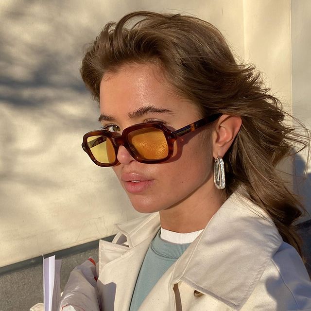 cabelo-castanho-claro Selfie de uma mulher. Ela usa uma blusa azul, casaco bege como sobreposição, brinco grande prata e óculos de sol com a armação dourada e lentes amarelas e cabelo solto. Ela olha para a câmera e não sorri.