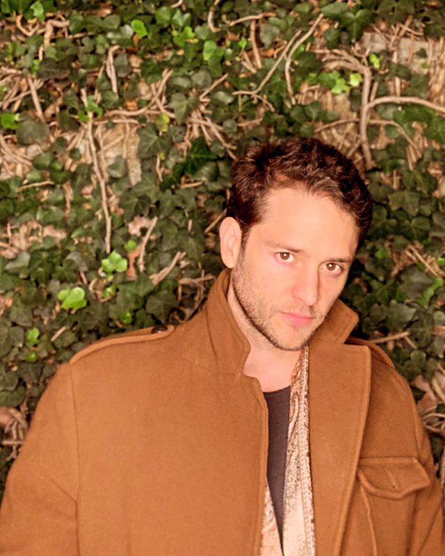 Foto de Christopher Uckermann do RBD com expressão neutra usando um casaco marrom e posando na frente de um fundo com folhas verdes