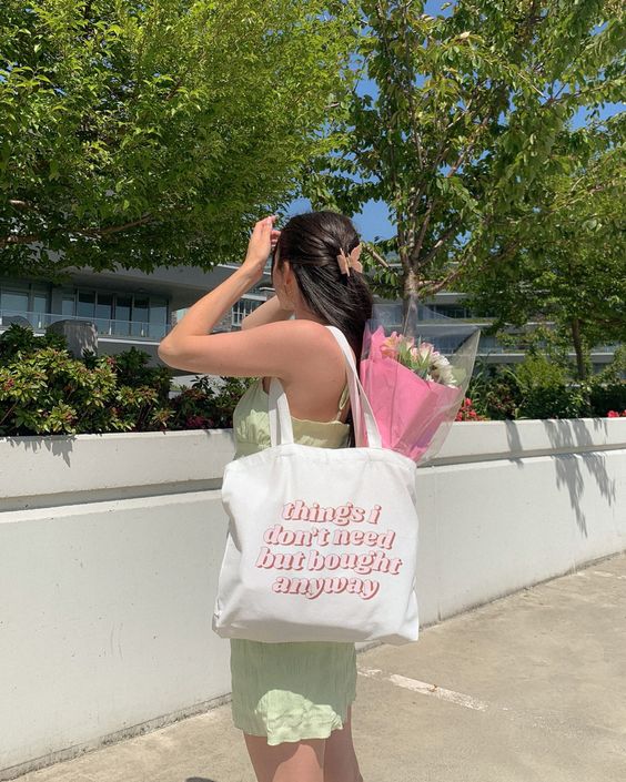ecobag-look Foto de uma mulher na rua. Ela usa um vestido verde menta, ecobag com estampa rosa claro com um buquê de flores dentro e cabelo preso com uma piranha. Ela está de lado e com a mão esquerda no cabelo, não olha para a câmera e não sorri.