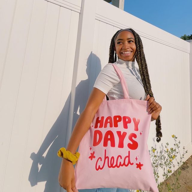 ecobag-rosa-look Foto de uma mulher em frente a um muro branco. Ela usa uma blusa de gola alta branca, calça jeans e ecobag rosa com