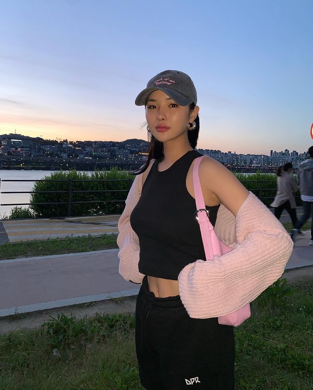 look-com-regata-preta Foto de uma mulher na rua. Ela usa um boné escuro, regata preta, casaco peludinho rosa como sobreposição, calça moletom e bolsa baguete rosa. Ela está com as mãos na cintura, olha para a foto e não sorri.