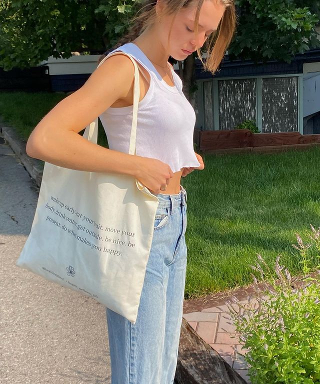 Foto de uma menina na calçada. Ela usa um top cropped branco, calça mom jeans e ecobag com estampa escrita preta e cabelo preso. Ela olha para o chão e está com as duas mãos segurando o top cropped.