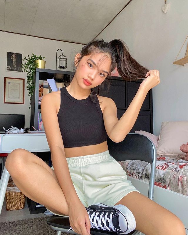 look-regata-preta-bermuda-moletom Foto de uma menina sentada em um banco em um quarto. Ela usa uma regata preta, bermuda de moletom clara, tênis de cano alto preto e cabelo preso em um rabo de cavalo. Ela olha para a câmera e não sorri.