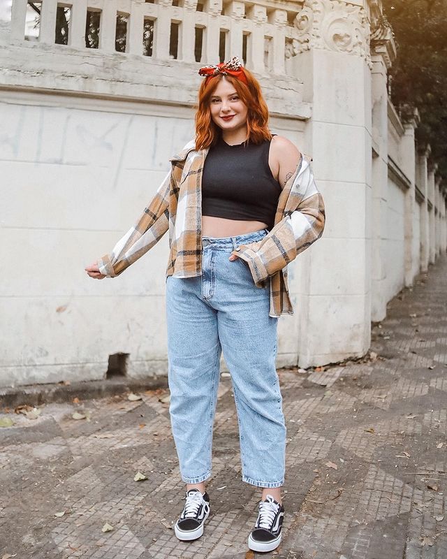 look-regata-preta-casaco-xadrez Foto de uma mulher em uma calçada. Ela usa uma regata cropped preta, casaco xadrez em tons de marrom, calça jeans, tênis preto e bandana no cabelo. Ela olha para a foto e sorri levemente.