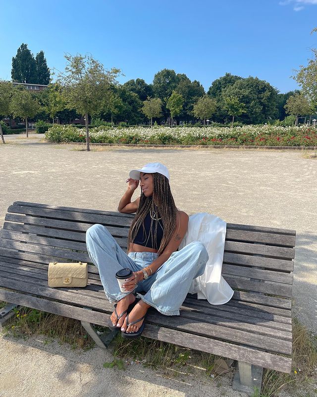 look-regata-preta-jeans Foto de uma mulher em uma praça sentada em um banco de madeira. Ela usa uma regata cropped preta, calça jeans oversized, chinelo, boné branco e bolsinha. Ela olha para o lado e segura um copo de café na mão esquerda.