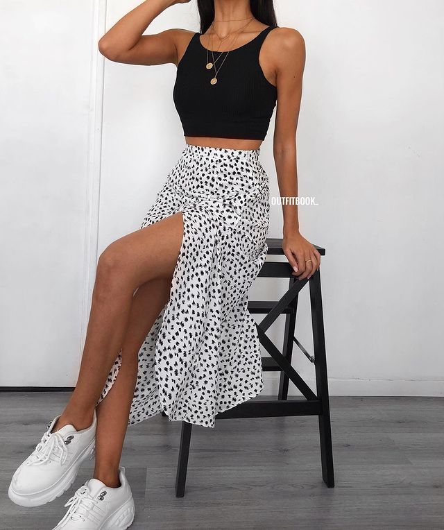 look-regata-preta-saia-midi Foto do look de uma mulher sentada em um banquinho preto. Ela usa uma regata preta, saia midi branca com estampa preta, tênis branco e colores dourados.
